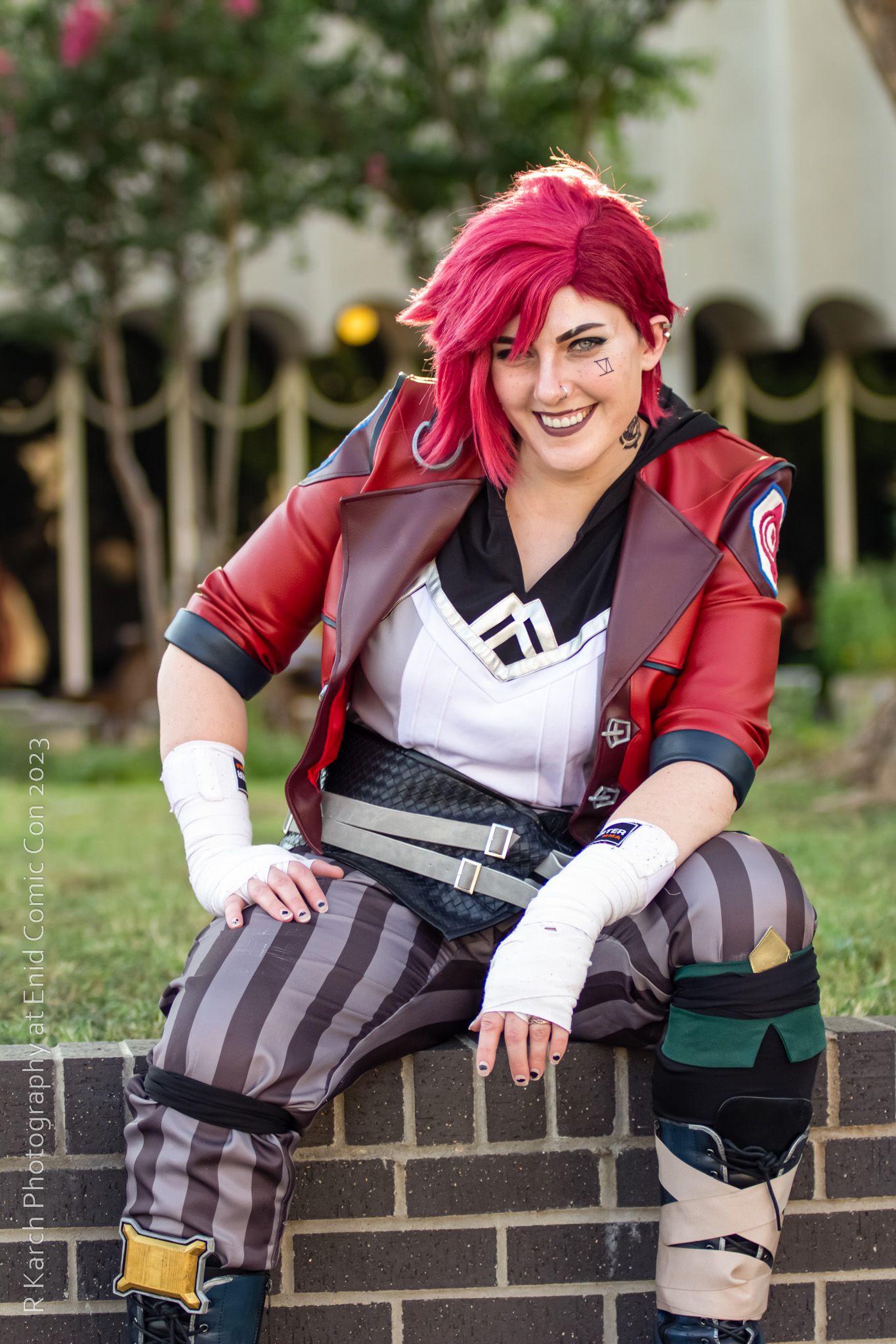 Cosplayer in red and black combat outfit with bright pink hair poses on brick wall outdoors.