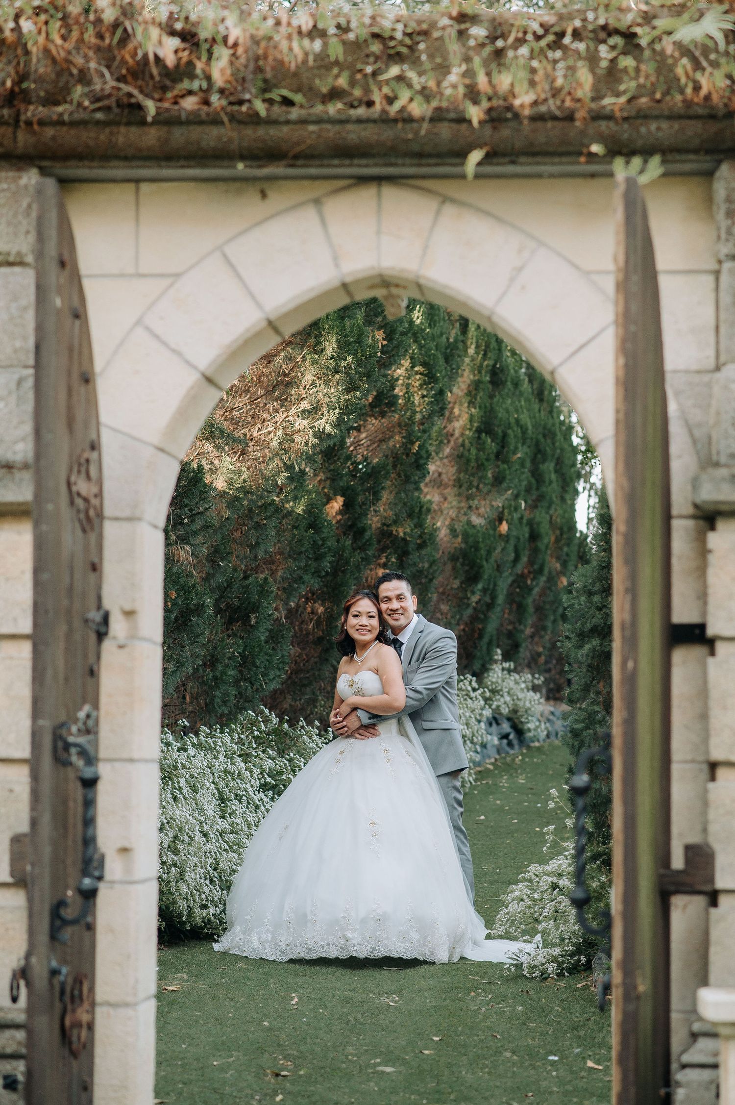A romantic wedding portrait framed by a stone archway surrounded by lush garden scenery.