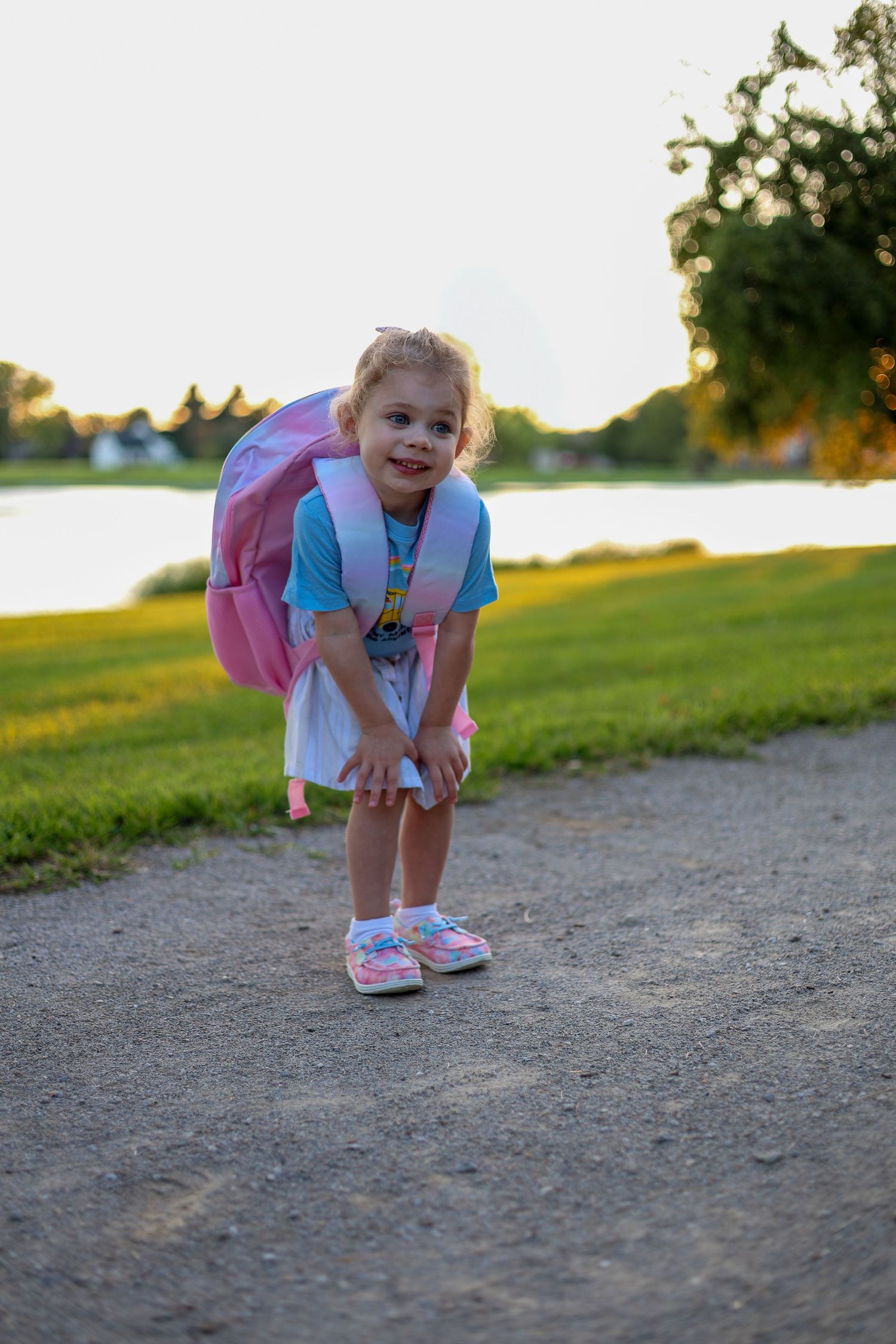 Child with pink backpack bends down on path near lake during sunset.