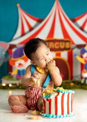 Happy toddler in red and white striped overalls enjoys birthday cake in front of circus-themed backdrop with red tents for cake smash photos