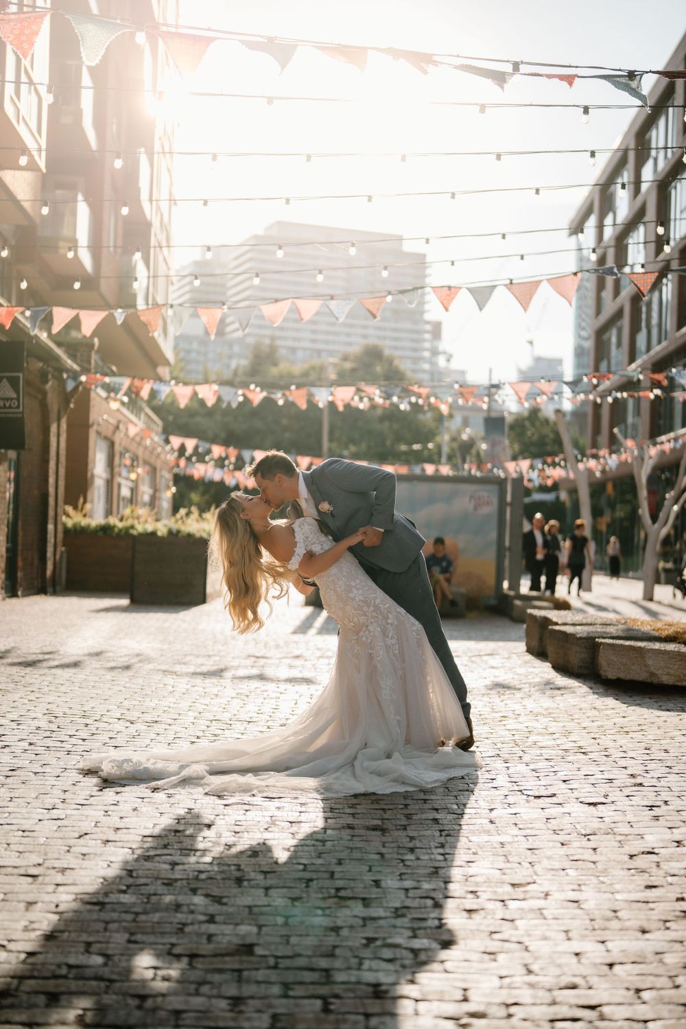 A romantic wedding couple shares a dip kiss on a cobblestone street decorated with string lights and bunting.