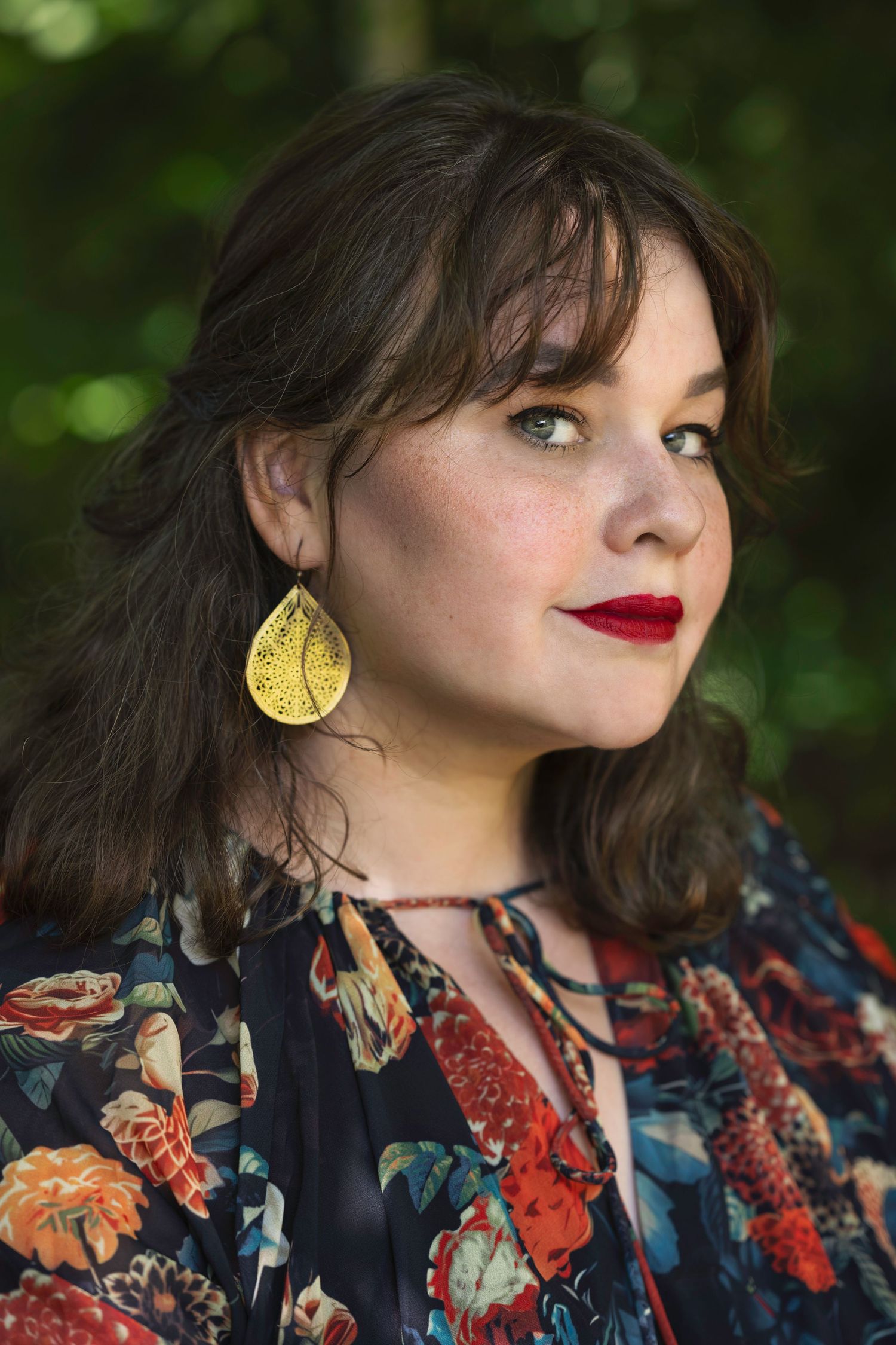 Woman with dark hair, red lips, and gold earrings wearing floral blouse poses outdoors in natural light.