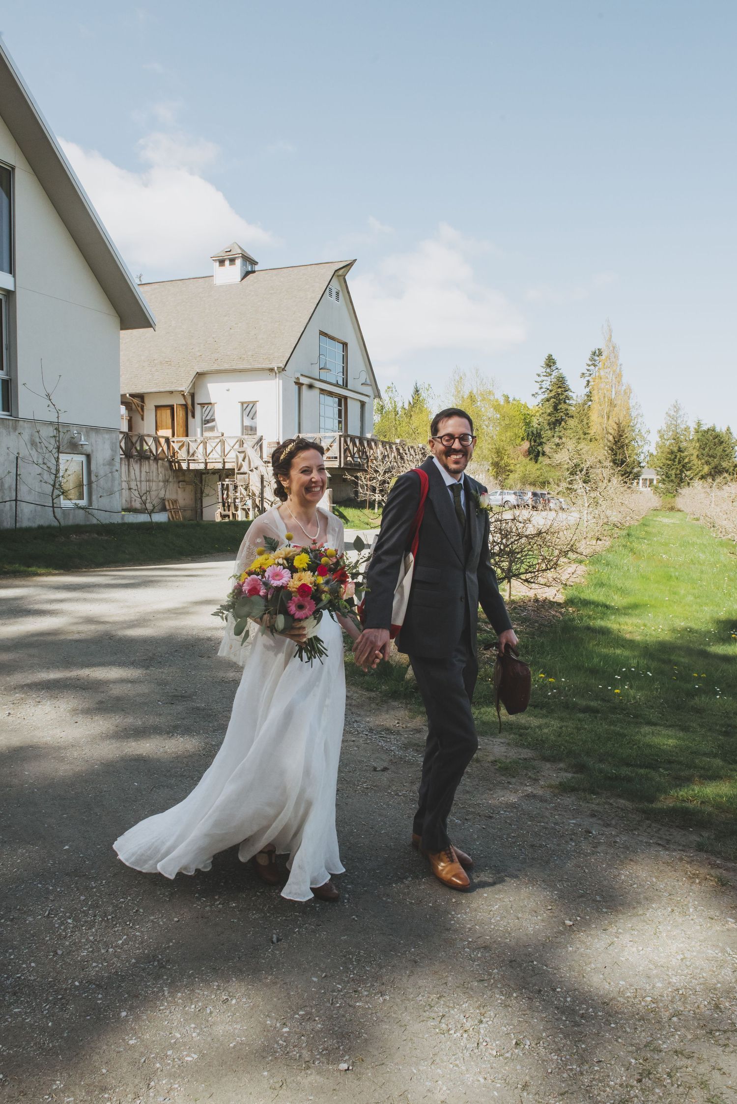 Newlyweds walk hand in hand past a white farmhouse carrying a colorful bouquet.