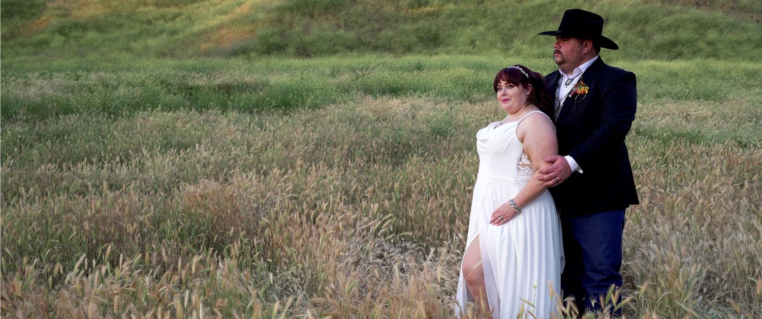 A couple in formal wedding attire walks together through a grassy field during sunset.