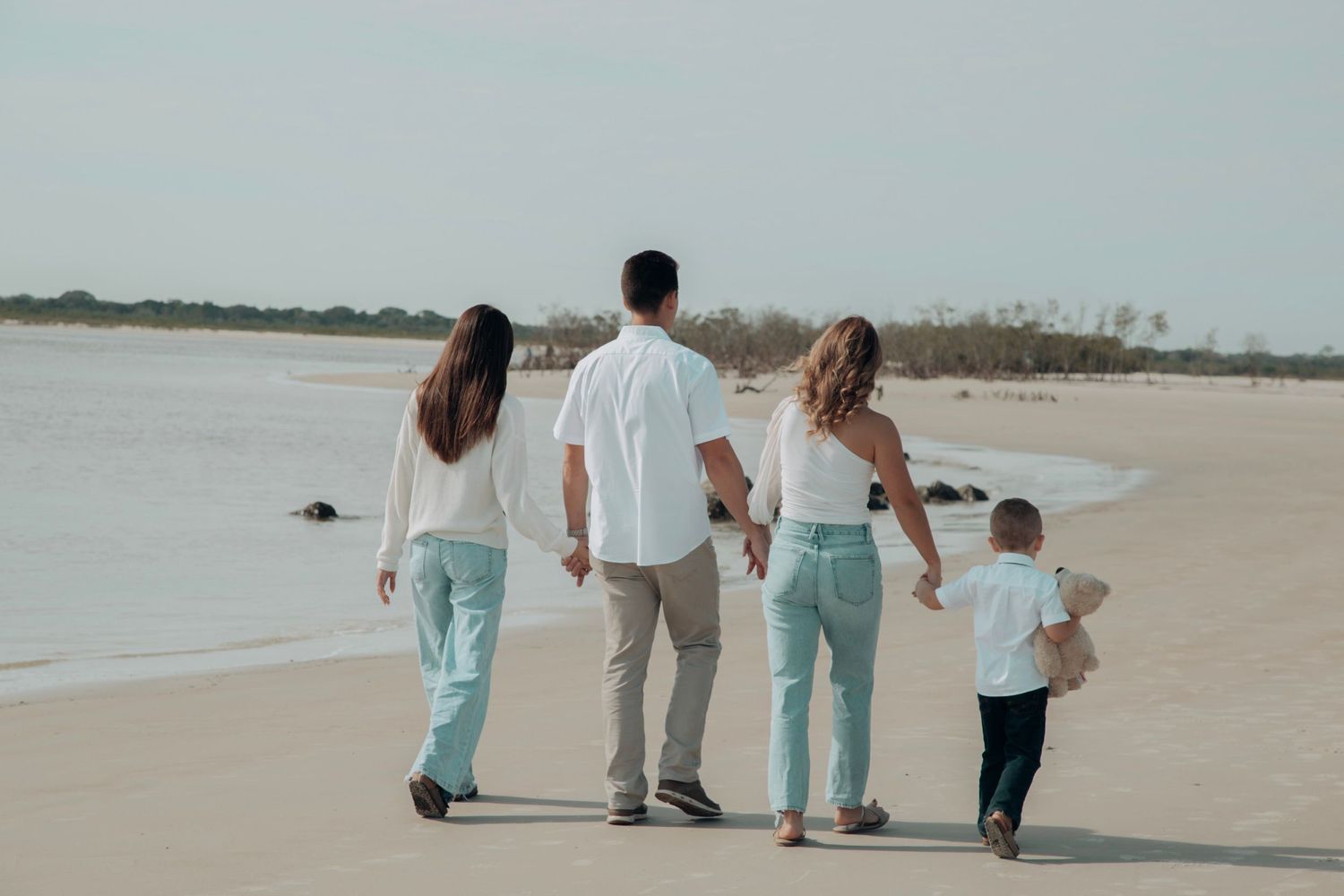 Family of four walks hand-in-hand along a serene sandy beach at sunset.