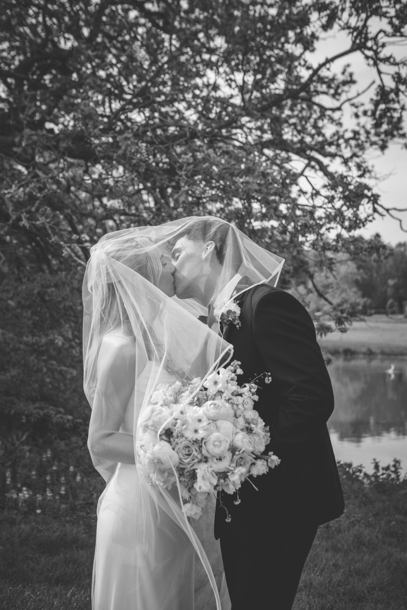Black and white photo of a bride and groom kissing under a veil outdoors, bride holding a bouquet near a lake.