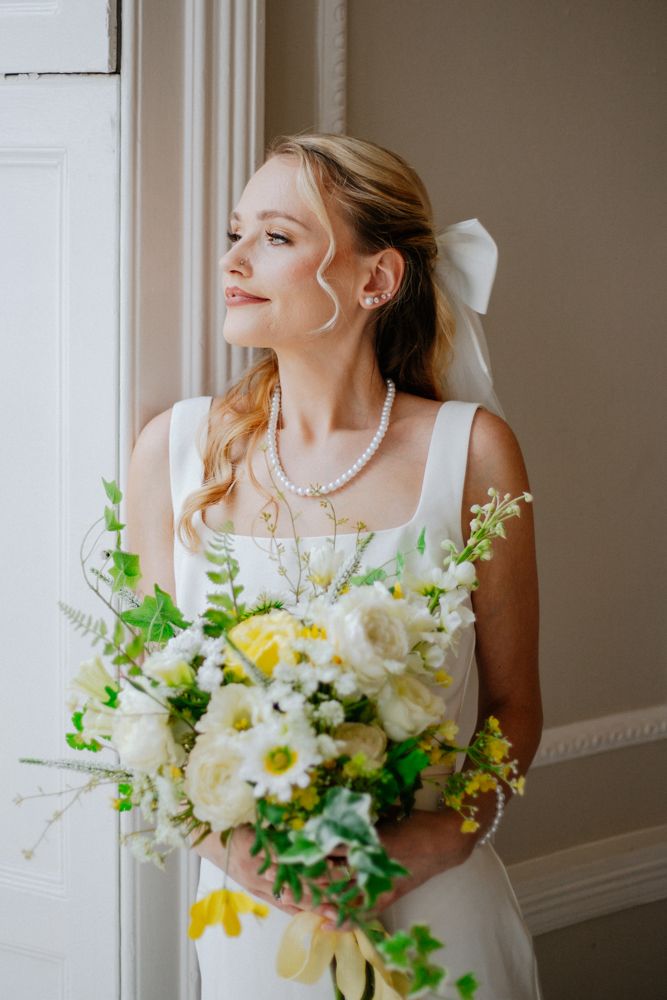 A bride holding a bridal bouquet features white daisies, yellow flowers and green foliage in a romantic spring arrangement looking left.
