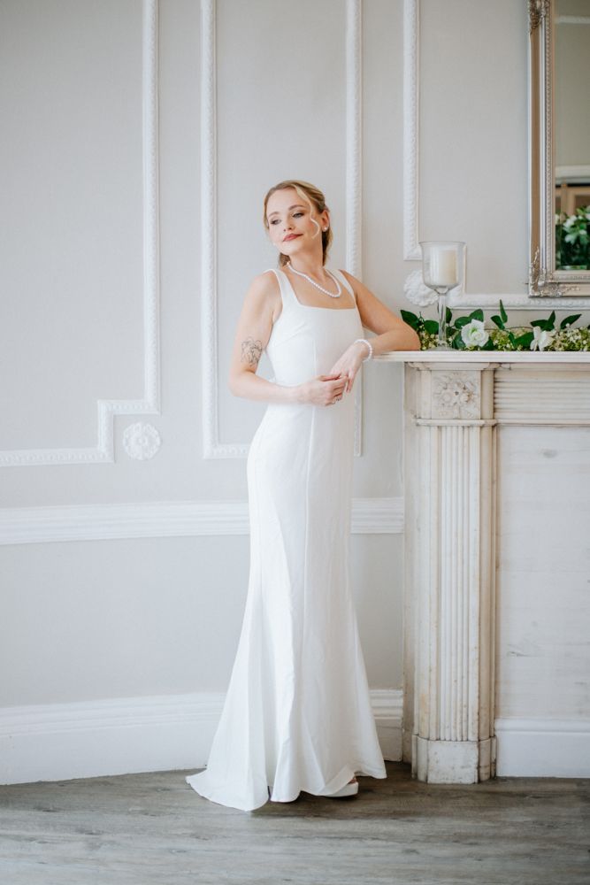 A bride in a white wedding dress stands gracefully in a bright, minimalist room with ornate white moldings and decor.