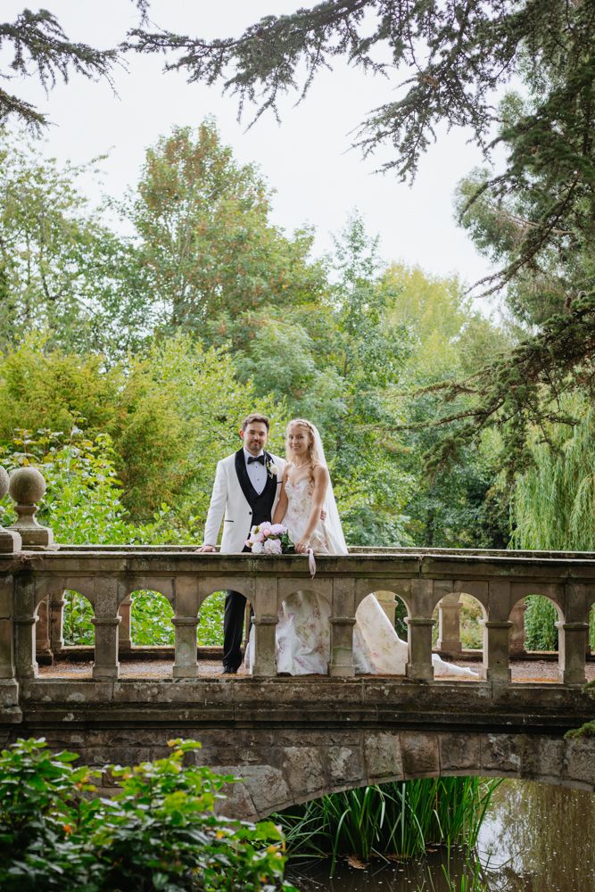 Bride and groom pose on a stone bridge at Henley Hall, Ludlow surrounded by lush greenery, bride holding a pink bouquet in a garden setting.