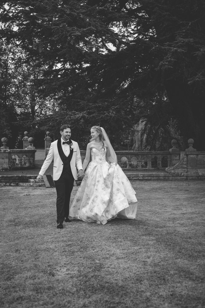 Black and white photo of a bride and groom walking hand in hand through Henley Halls elegant garden grounds on their wedding day.