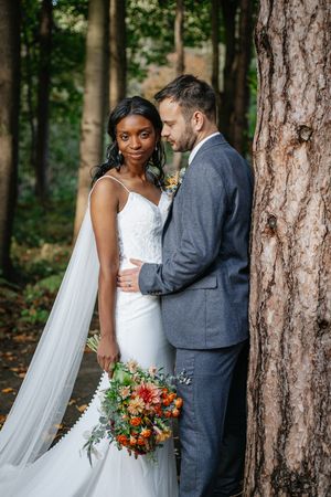 Romantic wedding couple embraces among tall pine trees in a forest setting with vibrant autumn bouquet.