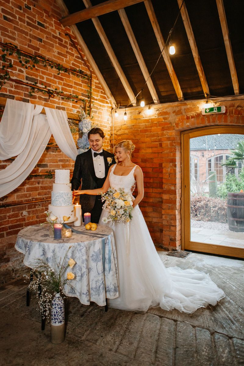Elegant wedding couple poses in rustic brick venue with flowing white dress and romantic floral arrangements.