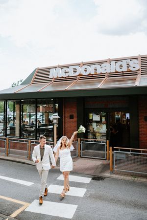 Newlywed couple in wedding attire holding hands crossing street in front of McDonald's restaurant.
