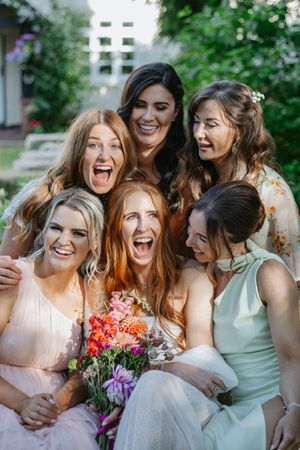 Bridesmaids in pastel dresses laugh together while holding colourful floral bouquet in outdoor garden setting.