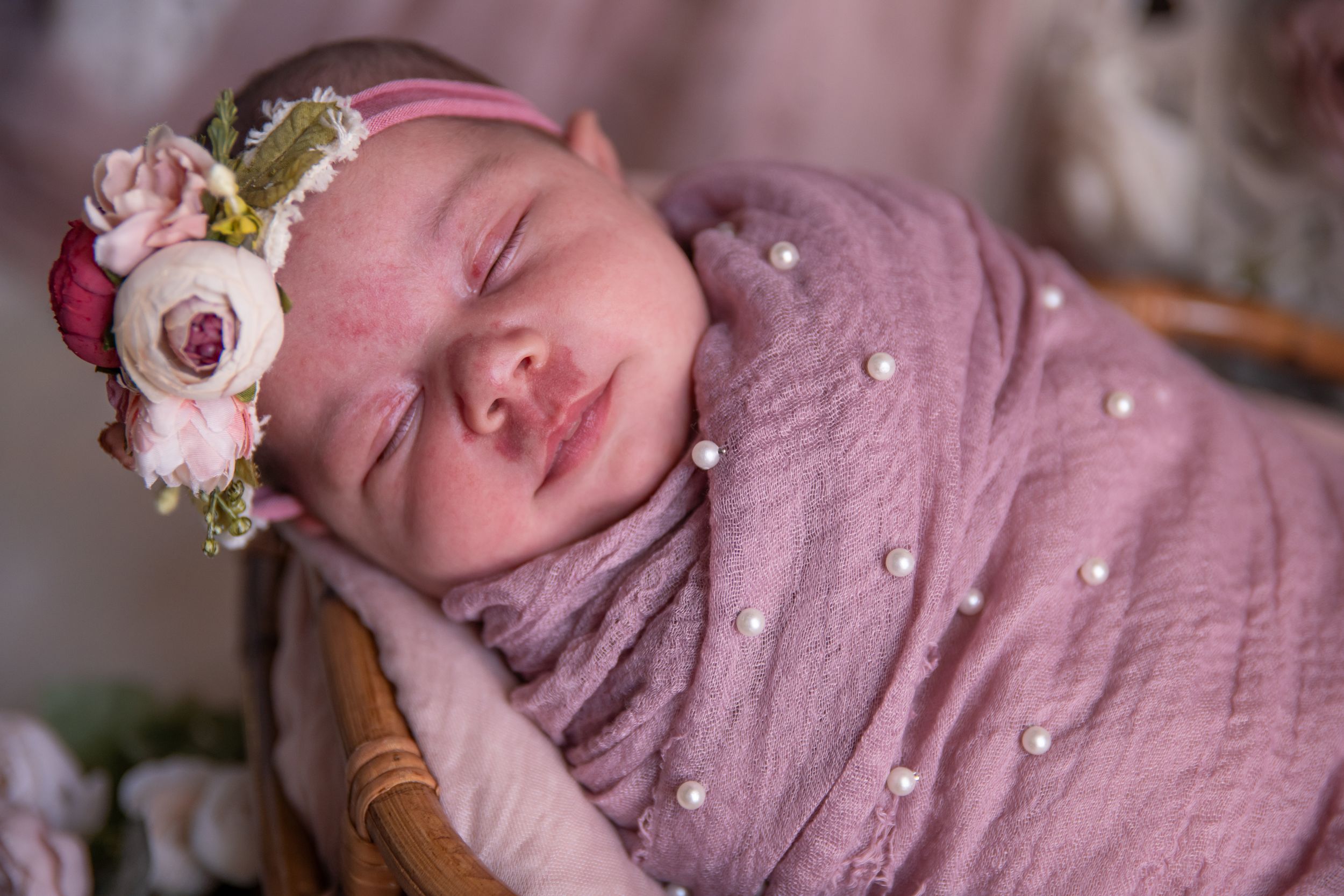 Sleeping newborn wearing pink wrap and floral headband rests peacefully in natural light.