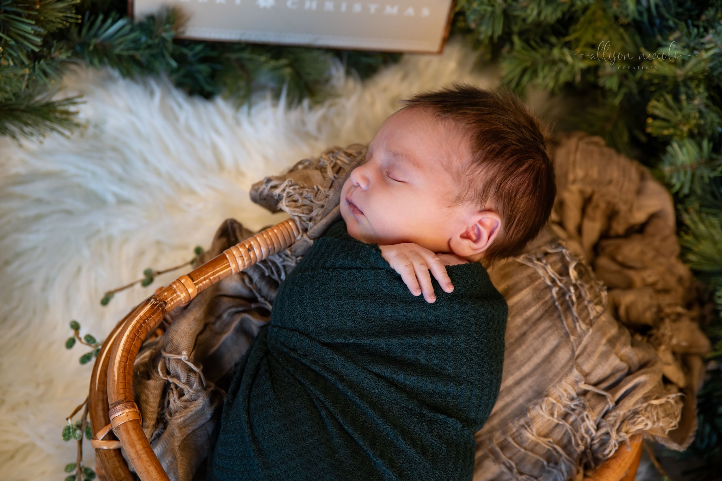 Baby sleeping peacefully in a wooden basket with cozy fur blankets and holiday greenery in the background.