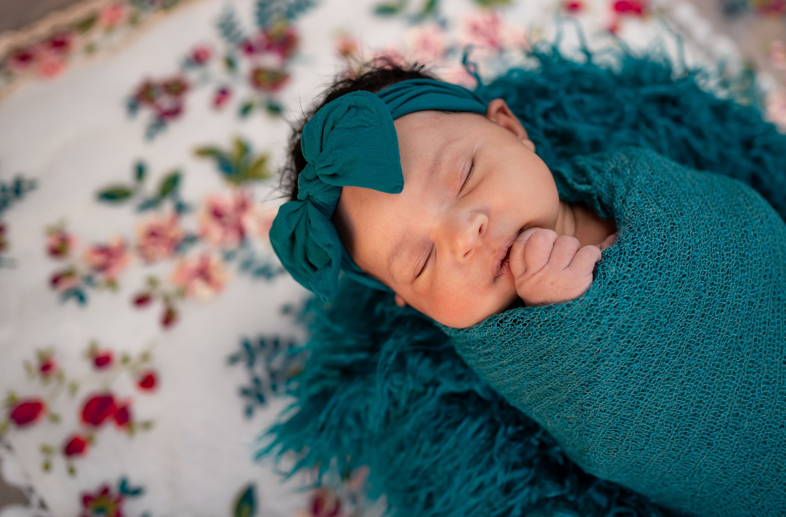 Sleeping newborn swaddled in teal knit wrap on floral blanket in soft lighting.