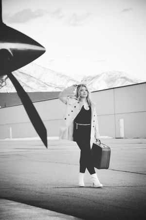 Black and white photo of a traveler with suitcase posing next to airplane propeller on airport tarmac with mountains behind.