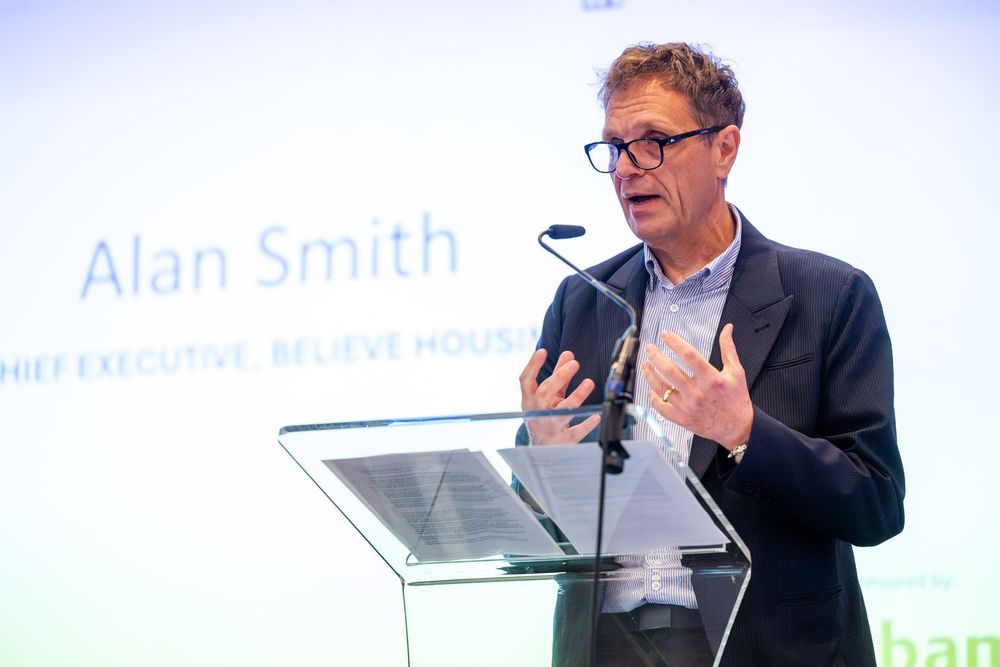 A speaker delivering a presentation at a podium during a professional event, gesturing while addressing the audience.
