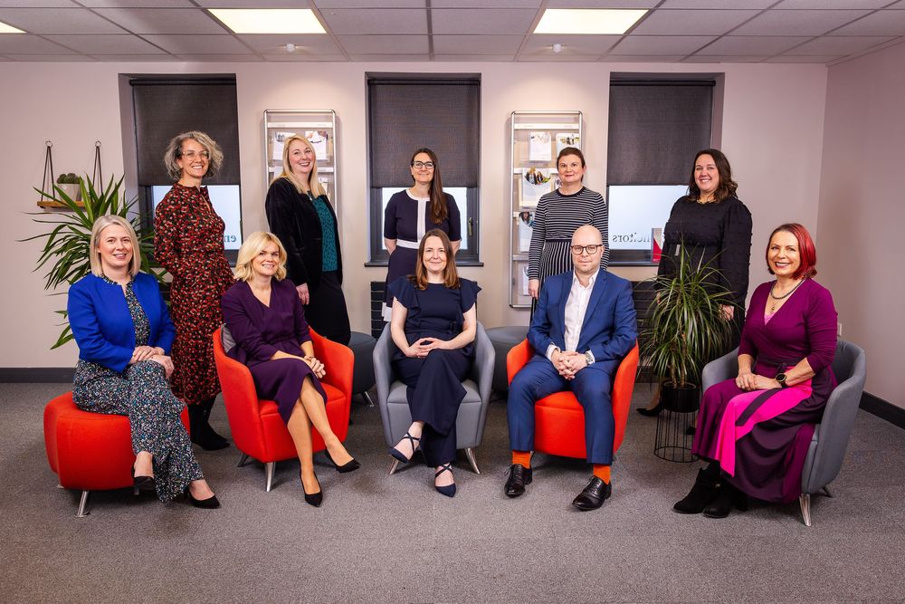Professional group portrait of ten colleagues seated and standing in an office setting with bright chairs and warm, modern decor.