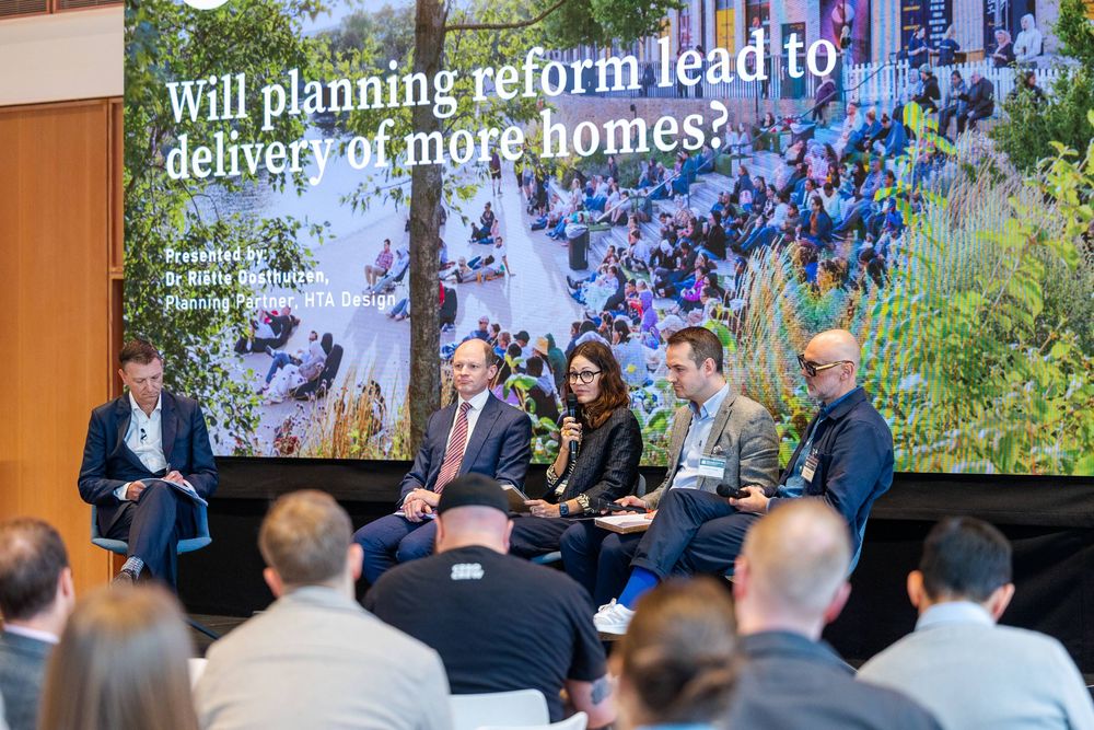 Panel discussion at a planning conference, with speakers on stage debating housing reform in front of a large presentation screen.