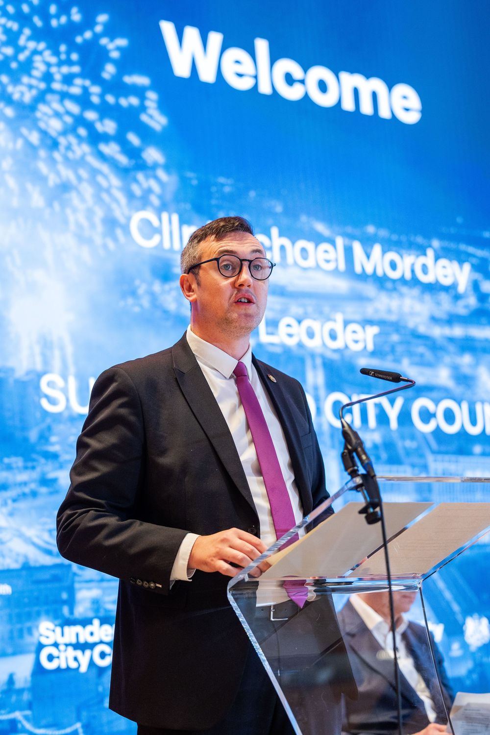 A speaker in a suit delivers a presentation at a business event, standing at a podium with a large welcome screen behind him.