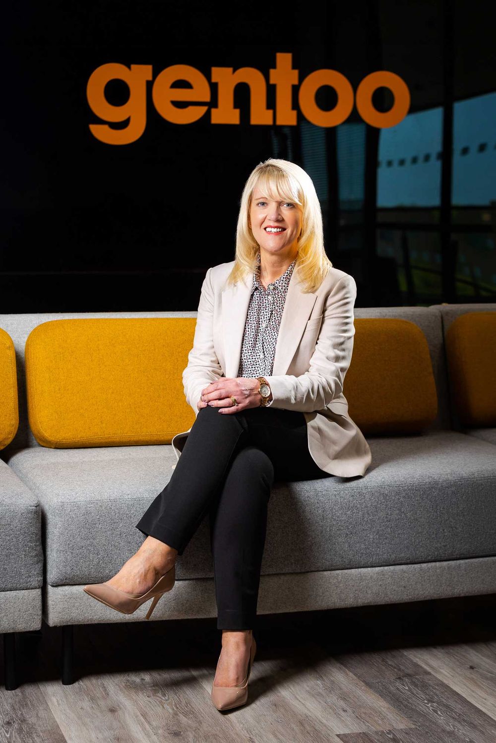 Professional corporate portrait of a female CEO seated on a modern sofa, with branded backdrop in a contemporary office setting.