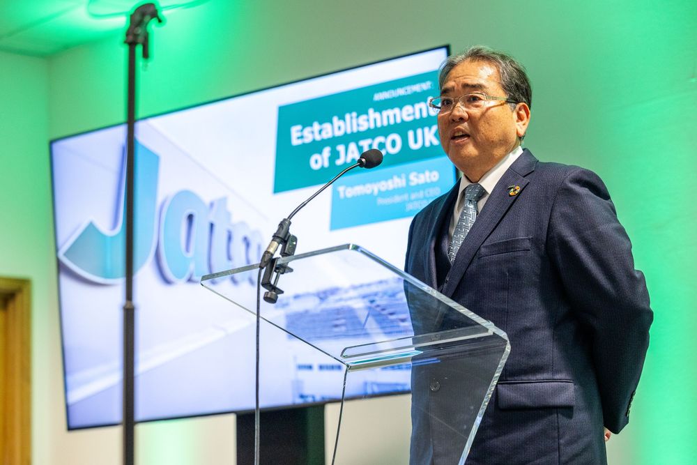 Business leader speaks at a podium during a corporate event, with a presentation screen behind him announcing the establishment of JATCO UK