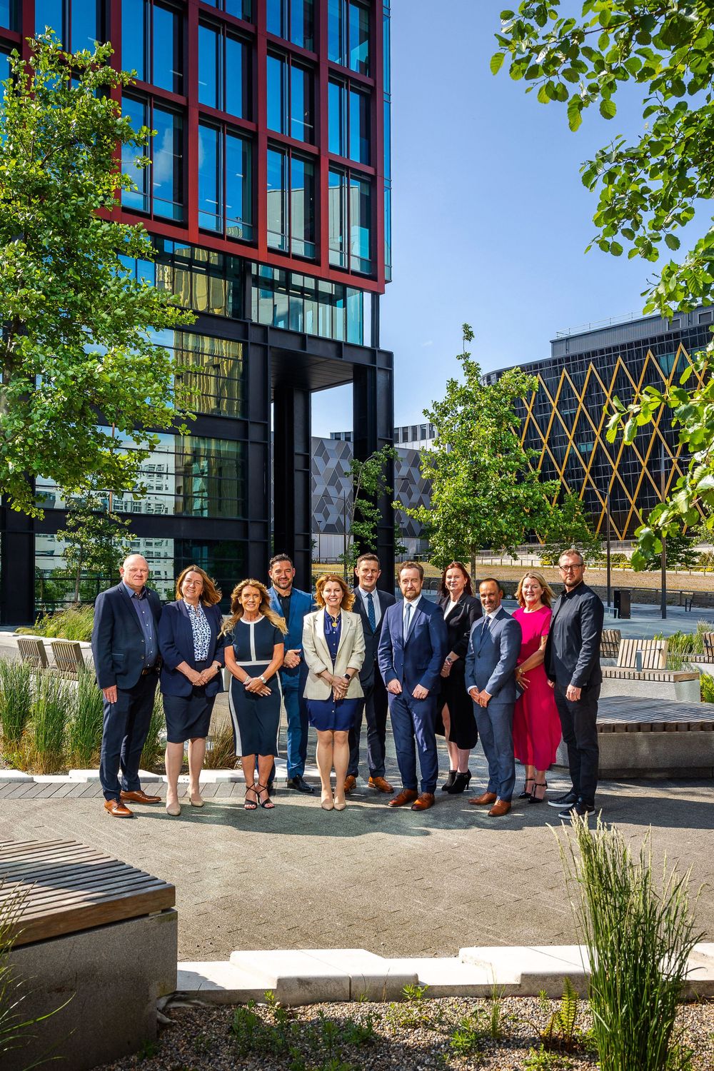 Corporate group portrait outdoors in a modern urban setting, with team members standing in front of contemporary office buildings.