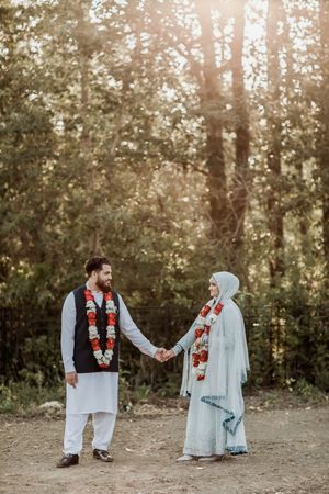 A couple in traditional South Asian wedding attire holds hands in a sunlit forest setting during a ceremony.