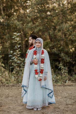 Couple in traditional Indian wedding attire adorned with red and white floral garlands embrace outdoors.
