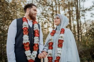Couple in formal attire wearing matching red and white floral garlands stand in a wooded setting during a ceremony.