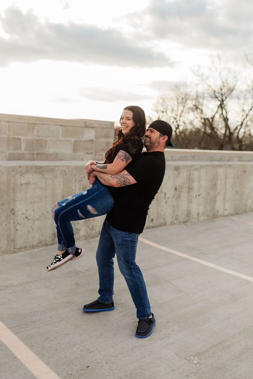 A man in black shirt and jeans lifts a smiling girl outdoors, sharing a joyful moment on a concrete terrace, in Indiana.