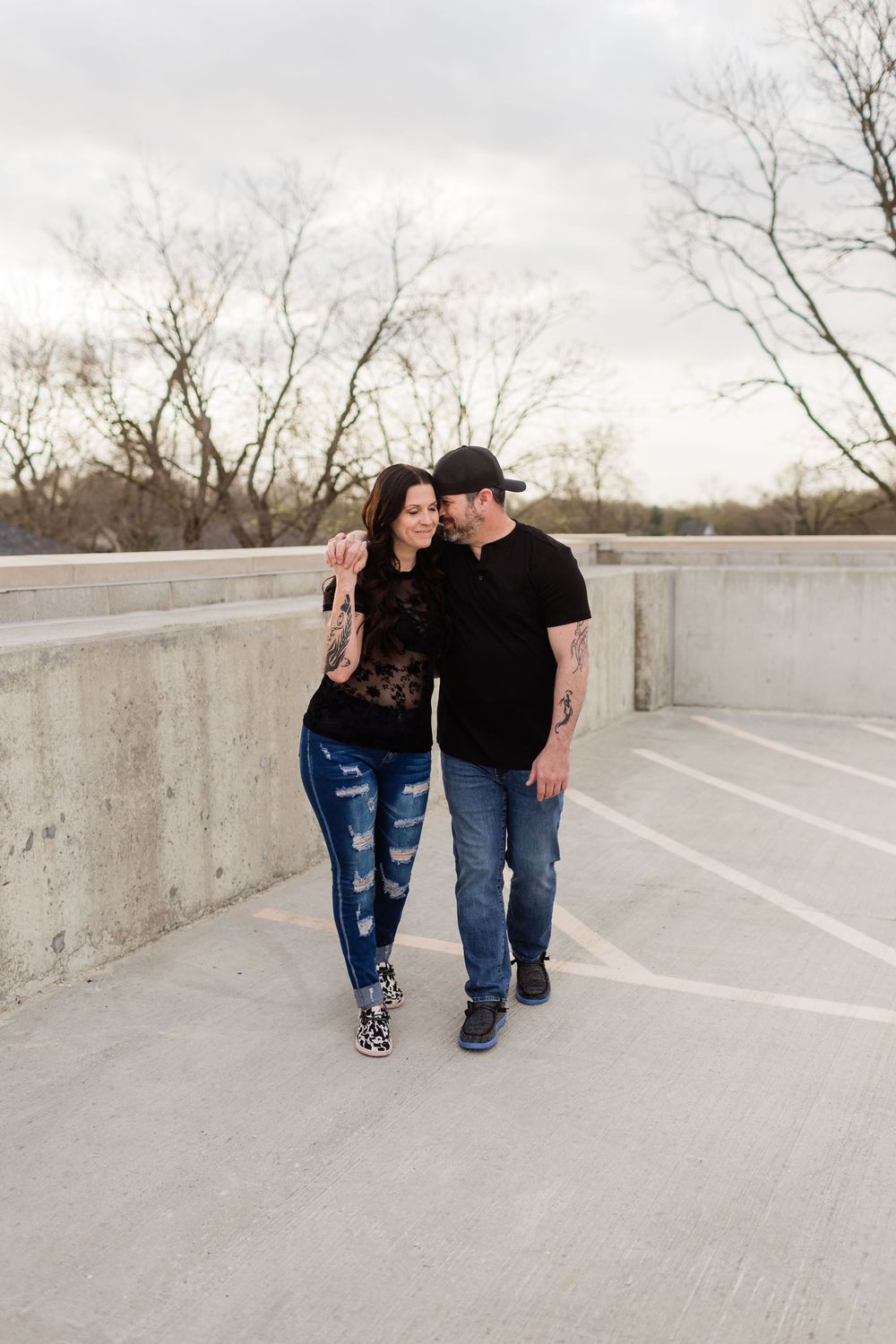 Couple in matching black tops and jeans walking closely together on an outdoor concrete terrace with bare trees, in Indiana.
