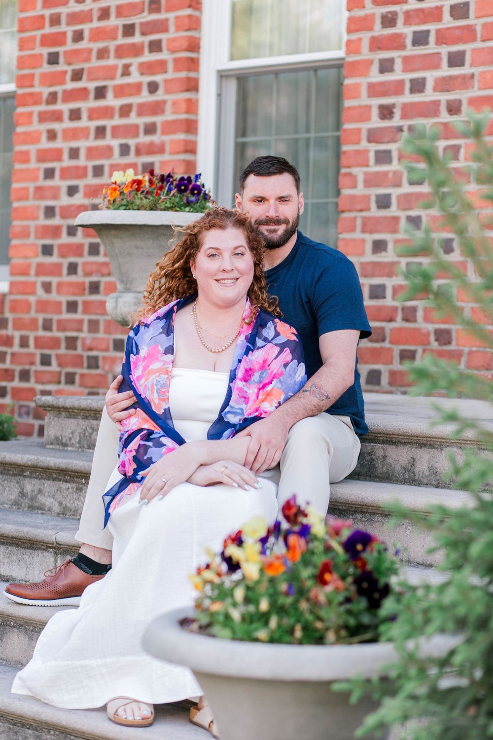 Couple sitting on brick building steps surrounded by colorful flower planters, woman in floral jacket and white dress, man in navy shirt.