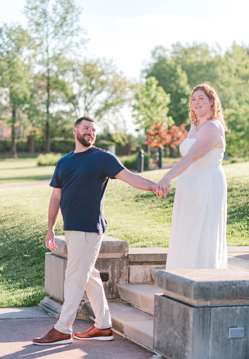 Couple holding hands on stone steps in a sunny park, woman in white dress and man in navy shirt smiling.