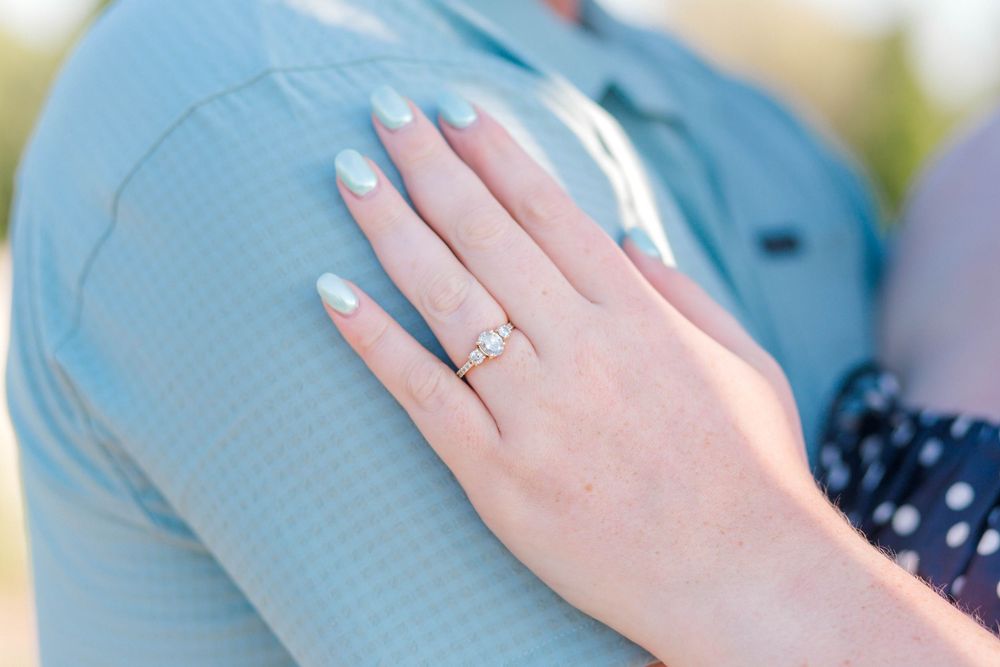 Close-up of a hand with mint nails wearing a delicate diamond engagement ring, resting on a light blue shirt.