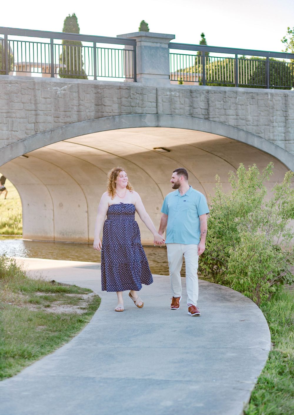 Couple holding hands walking on a curved path under a stone bridge arch surrounded by lush greenery.
