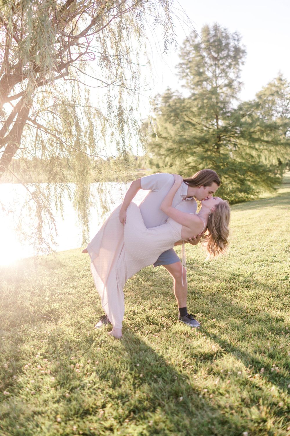 Couple sharing a romantic dip kiss in golden sunlight by a lake, surrounded by lush green grass and willow trees.