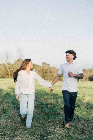 A couple holds hands and walks through a green field, smiling at each other on a sunny day.