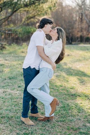 A couple sharing a romantic kiss outdoors on green grass, wearing white tops and jeans, with trees in the background.