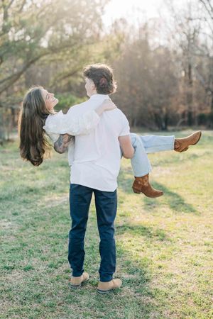 Man carrying woman in his arms outdoors, both in white tops and jeans, sharing a joyful moment in a sunlit park.