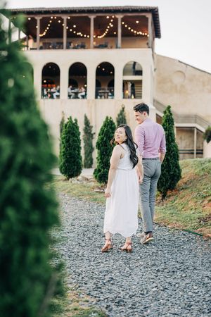 Couple walking on gravel path near elegant venue with string lights, woman in white dress looks back smiling.
