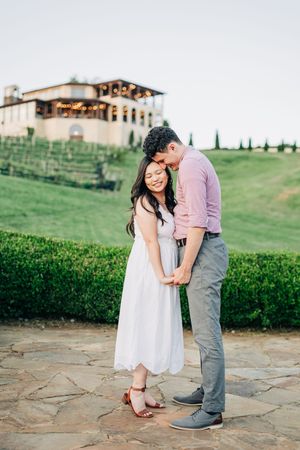 Couple holding hands and embracing on stone patio at vineyard estate during golden hour engagement session.