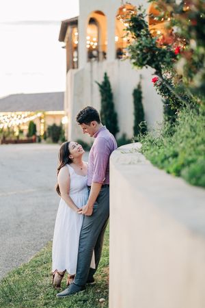 Couple smiling at each other while standing close together beside a wall with string lights at winery.
