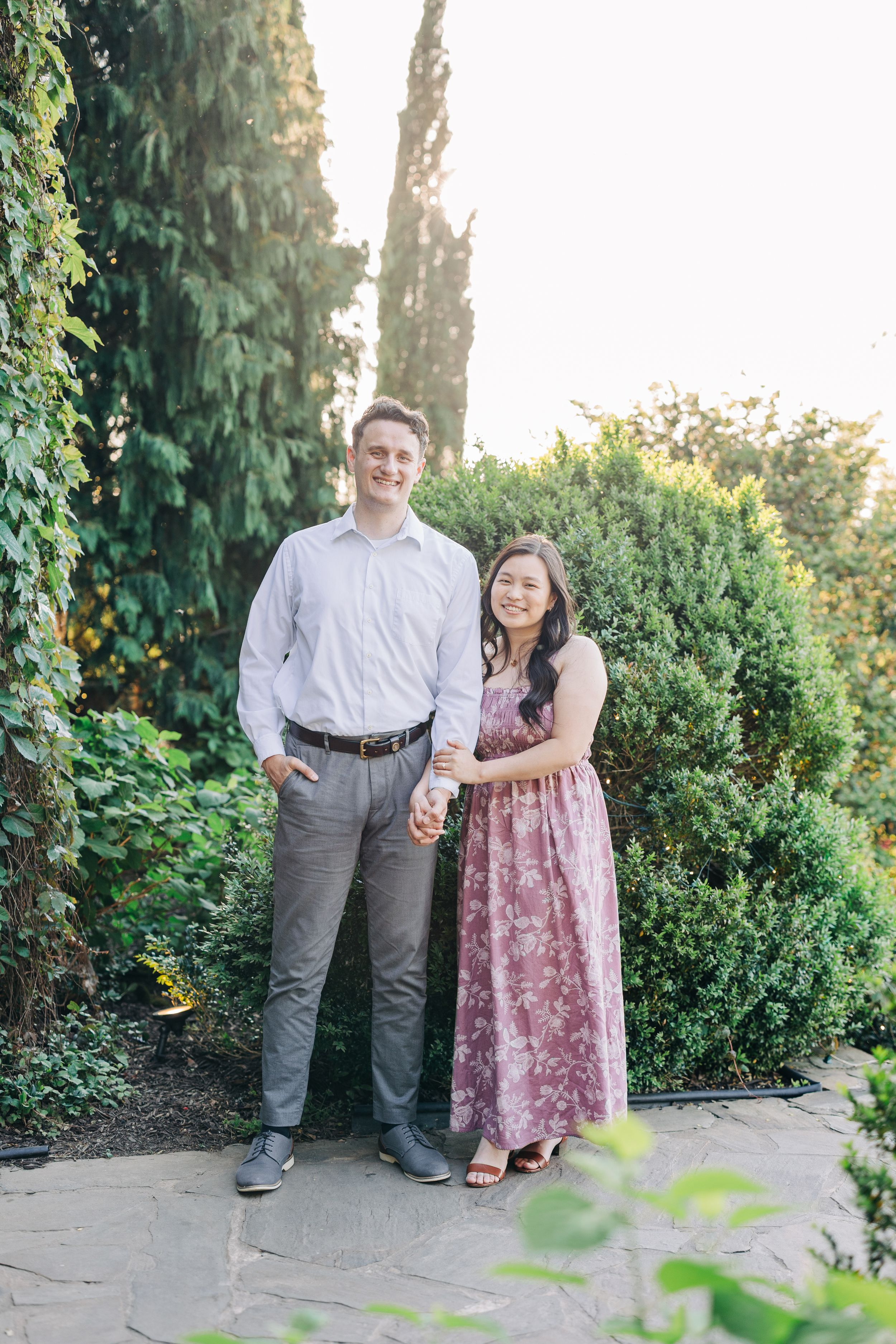 Couple holding hands smiling in lush garden, man in gray pants white shirt, woman in pink floral maxi dress.