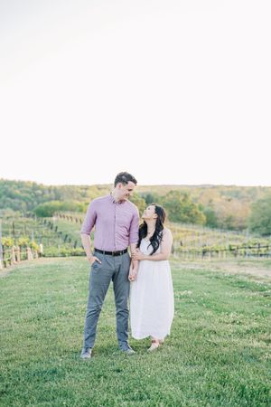 Couple sharing a romantic moment in a vineyard, woman in white dress leaning on man in pink shirt at golden hour.