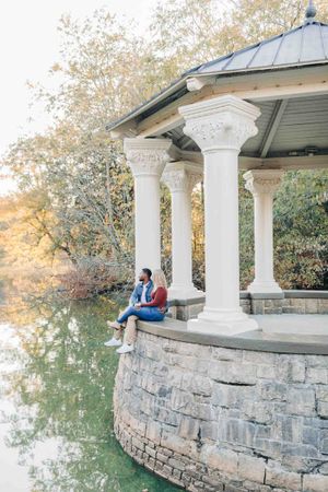 Couple sitting together at a elegant white gazebo by a reflective pond surrounded by autumn foliage.
