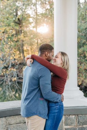 Interracial couple embracing and smiling at each other outdoors with sunlight and autumn trees in background.
