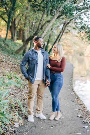 A happy interracial couple holds hands on a scenic park path surrounded by autumn trees near a calm river.